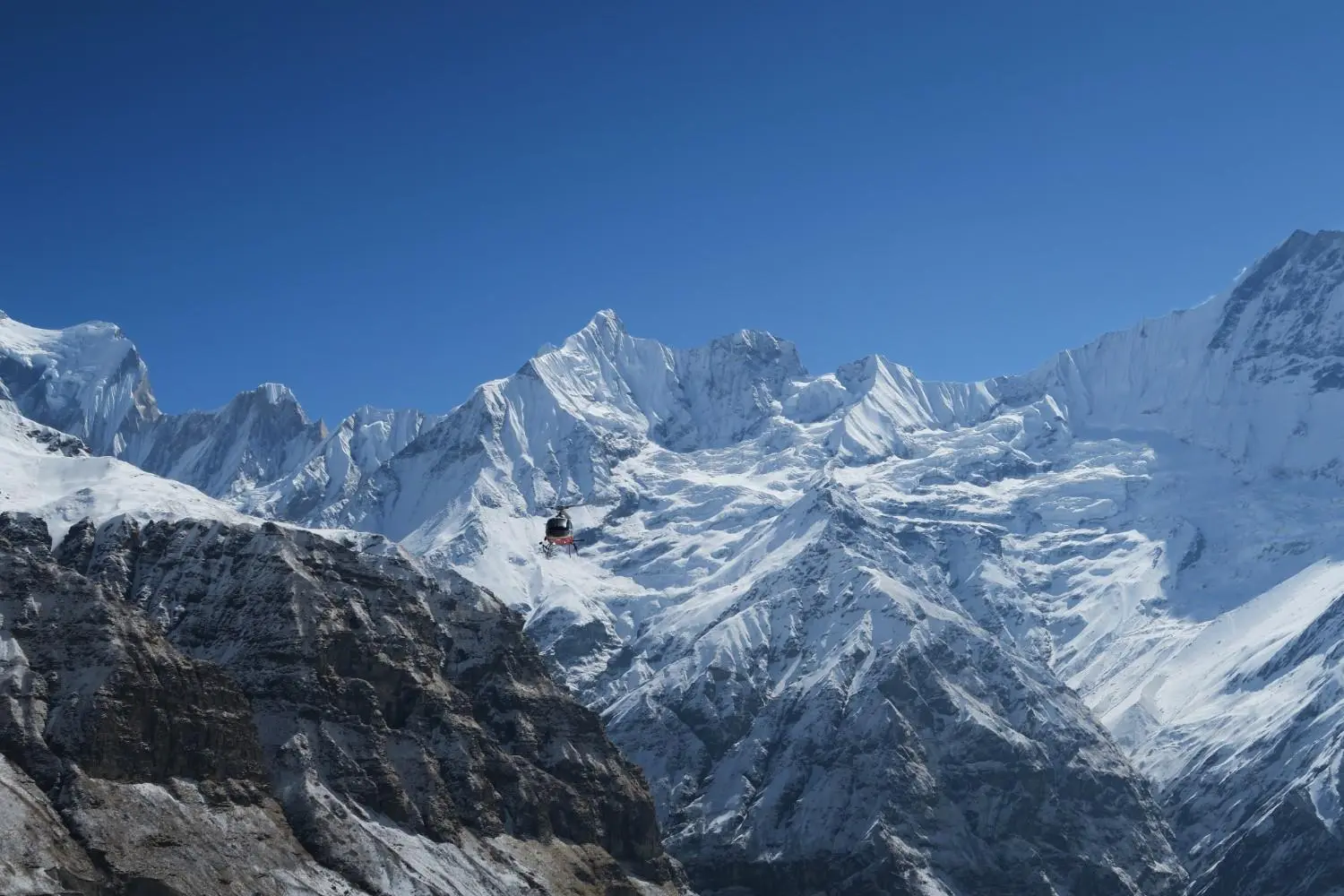 A small, dark helicopter flies in front of a giant, jagged Annapurna range covered in snow.