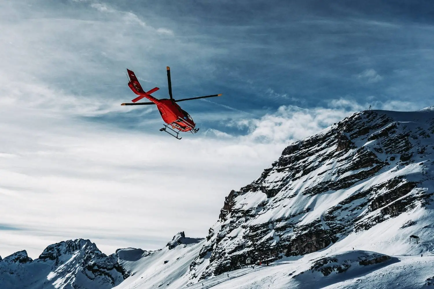 A red helicopter flies near a large, snow-covered mountain under a partly cloudy sky