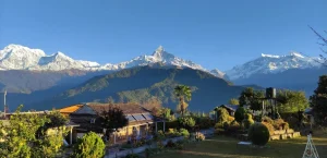 Panoramic view of Astam Village with traditional houses, lush gardens, and the snow-capped Annapurna mountain range under a clear blue sky in Nepal.