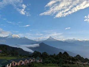 Scenic view from Australian Base Camp featuring mountain lodges, rolling hills, low-lying clouds, and the snow-capped Annapurna mountain range under a blue sky in Nepal.